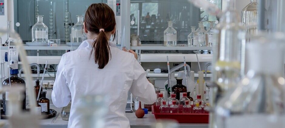Fotografia de uma cientista de costas em um laboratório trabalhando em uma bancada e analisando testes para covid-19 em Minas Gerais.